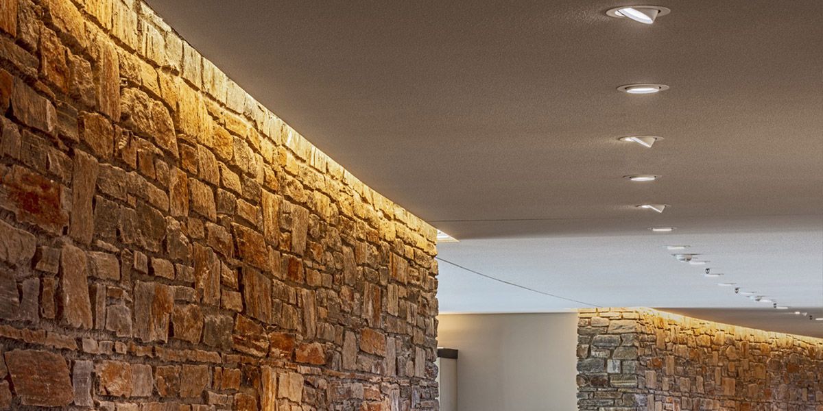 Natursteinwand mit indirekter, warmer LED-Beleuchtung und mehreren eingelassenen Deckenstrahlern in einem Gastronomiebereich.